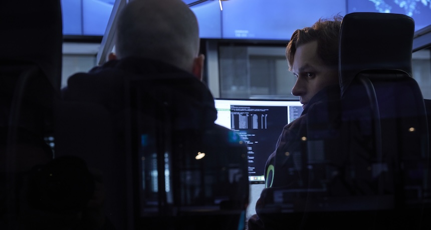 Two people sit in front of computer monitors displaying code in a dimly lit room, with reflections visible on the glass around them.
