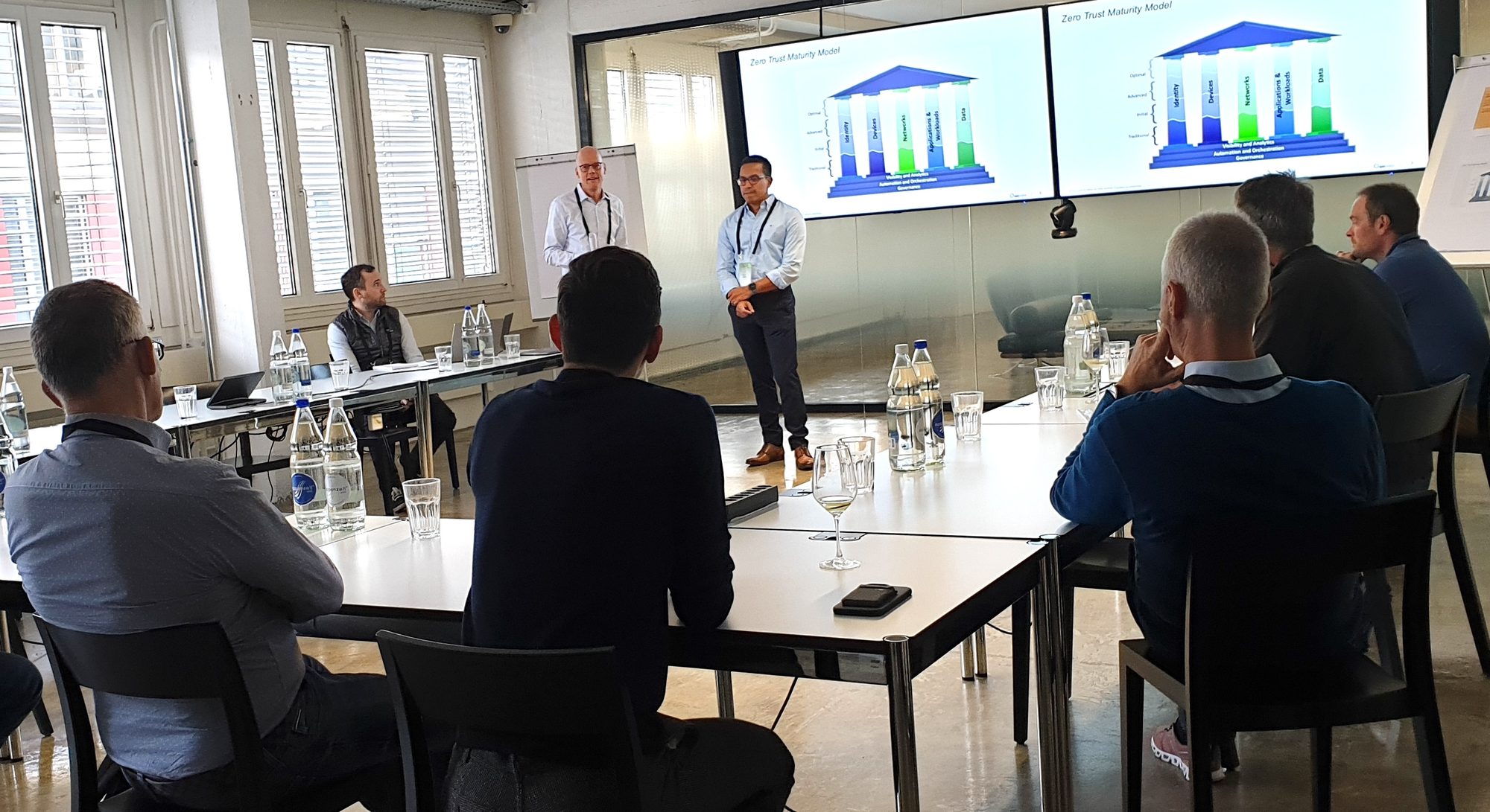 A group of people sit around tables in a bright conference room, listening to two men presenting graphs displayed on large screens at the front of the room. Bottles of water and glasses are on the tables.