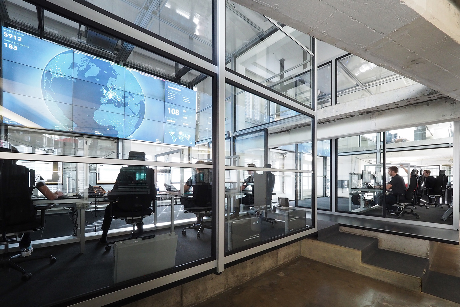 Office space with multiple workstations separated by glass walls. People are working at desks with computers. Large screen displays world map and data on the wall. The modern, industrial-style interior has exposed concrete and metal elements.