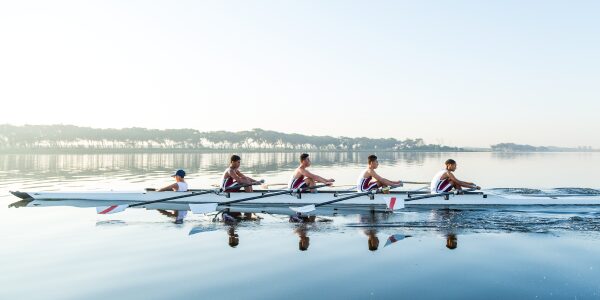 Fünf Menschen in einheitlicher rot-weisser Sportkleidung rudern in einem schmalen Boot über einen ruhigen See. Das klare Wasser spiegelt ihre Bewegungen. In der Ferne zeichnet sich unter dem wolkenlosen Himmel eine Baumreihe ab.