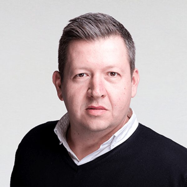 A man with short, dark hair and fair skin is wearing a black sweater over a white collared shirt. He is facing the camera with a neutral expression, standing against a plain, light grey background. His demeanor subtly prompts the question: what is SASE?