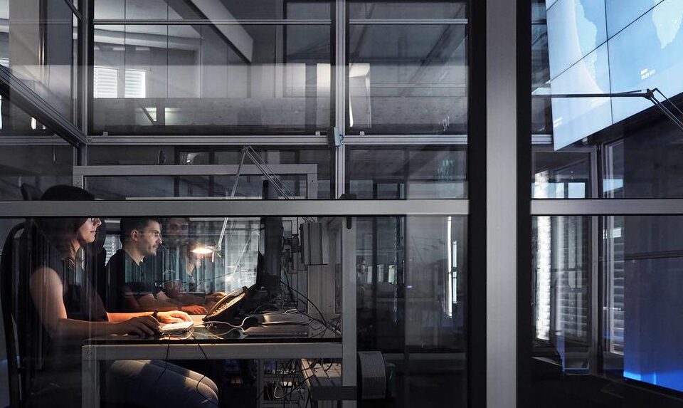 Several people are working at computer desks in a modern, glass-walled office environment. The room is dimly lit, with illuminated screens casting a blue glow. The setting appears to be focused on technology or software development.