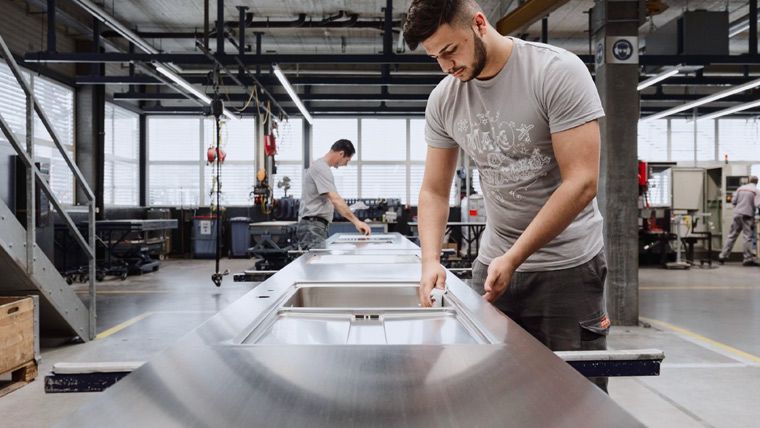 Two men work in a modern, well-lit factory on a production line assembling stainless steel components. The man in the foreground focuses intently on his task, while another man in the background works at a similar station. As they operate seamlessly like an SD-WAN backbone, both wear casual work attire.