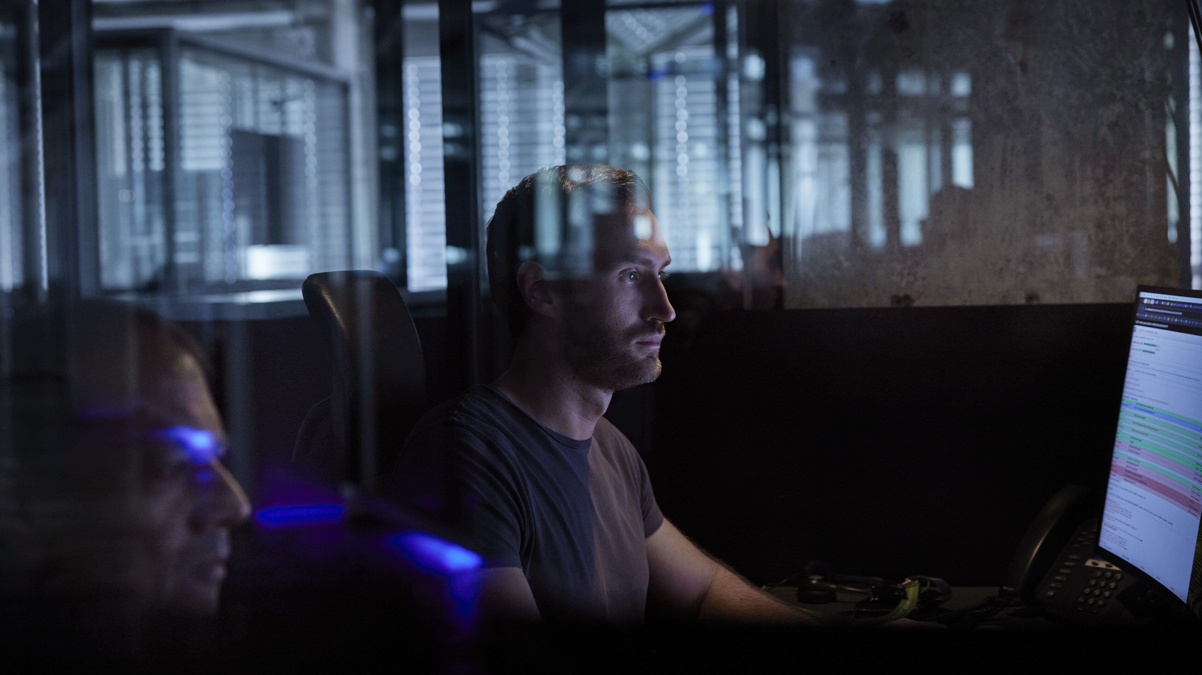 Une personne portant un t-shirt sombre est assise devant un écran d'ordinateur dans un bureau faiblement éclairé, concentrée. L'atmosphère est réfléchissante avec des surfaces vitrées, montrant diverses lumières et reflets en arrière-plan.