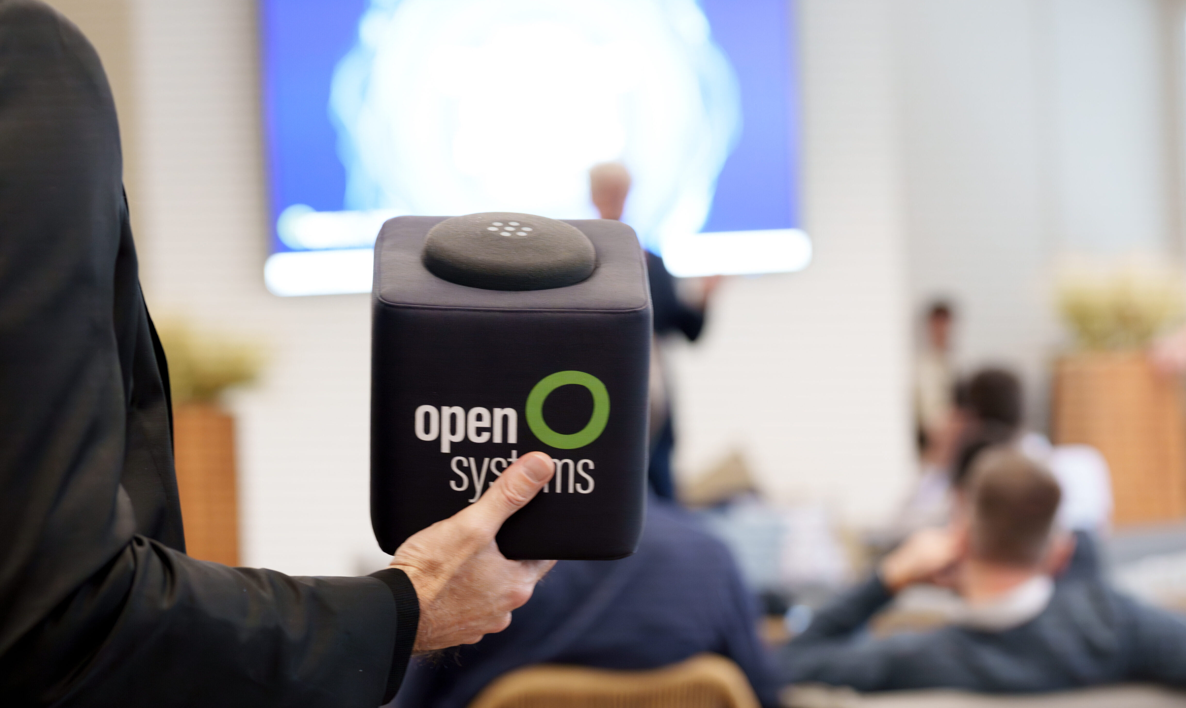 A person holds a cube-shaped object with "open systems" written on it in a conference room. In the background, people sit facing a large screen displaying a blurred presentation.