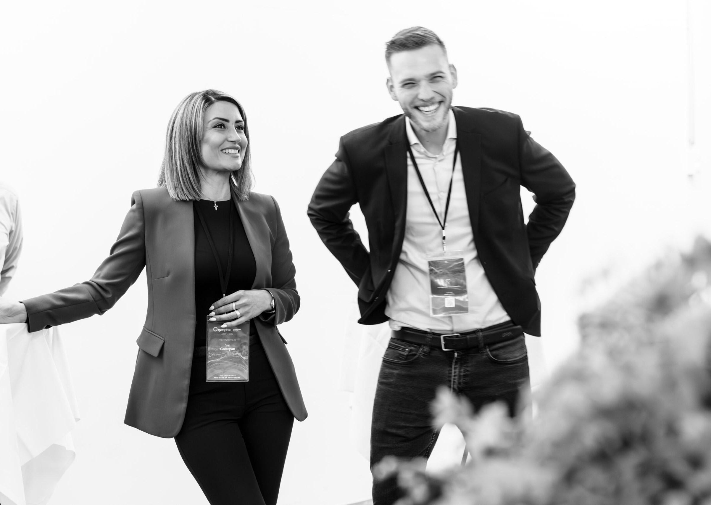 A black and white photo of two people, a woman and a man, in business attire, smiling and standing casually. Both have lanyards around their necks. They appear to be in a relaxed, social setting. The background is softly blurred.