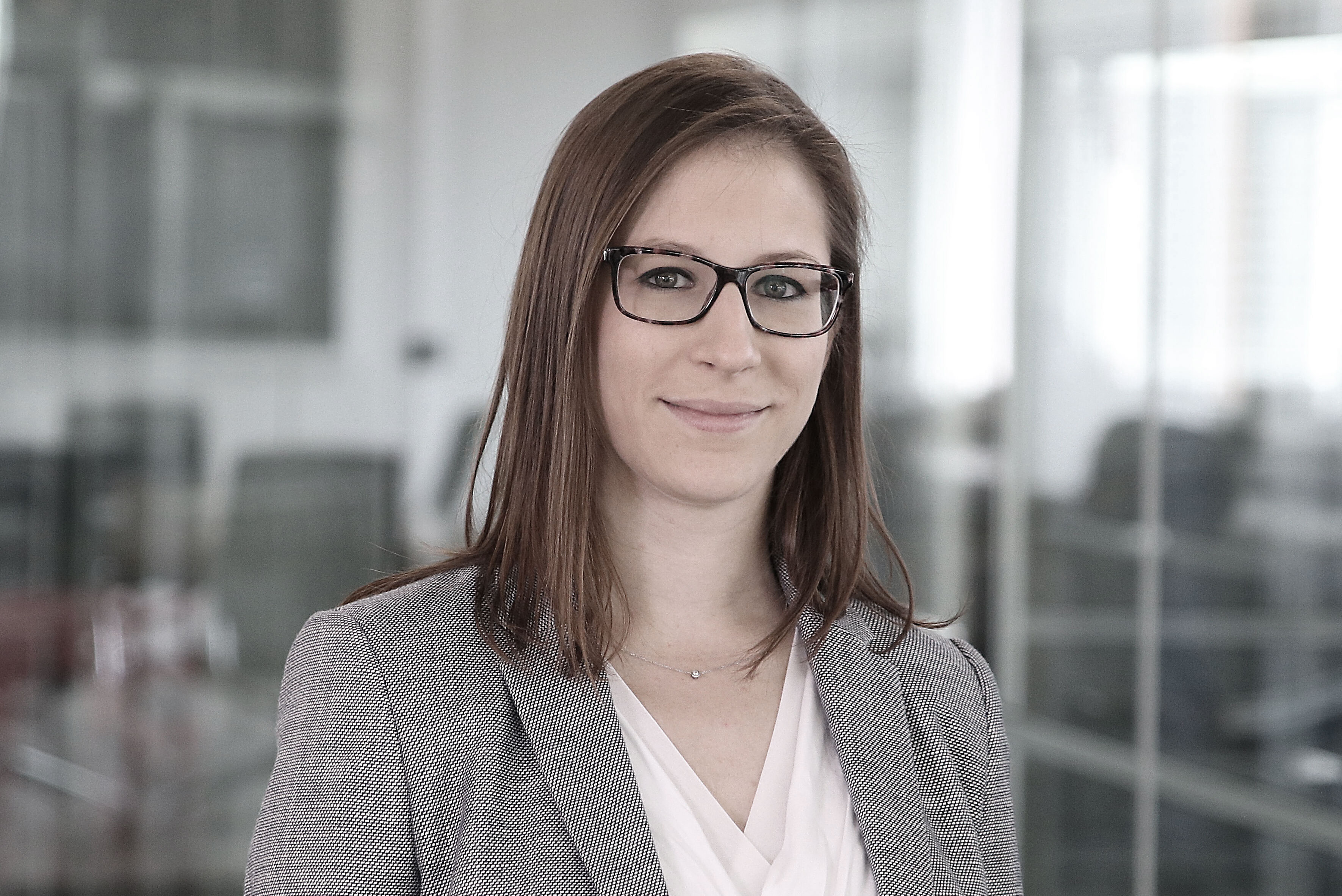 Stéphanie Falbriard, a woman with long brown hair and glasses, stands in a modern office with glass walls, wearing a gray blazer and white blouse. She is smiling slightly, while the background remains softly blurred.